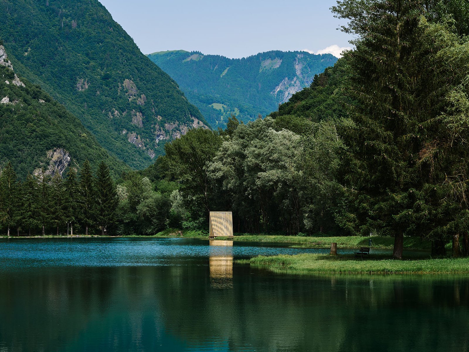 Timber architectural installation on the edge of a mountain lake, set within a forested alpine landscape.