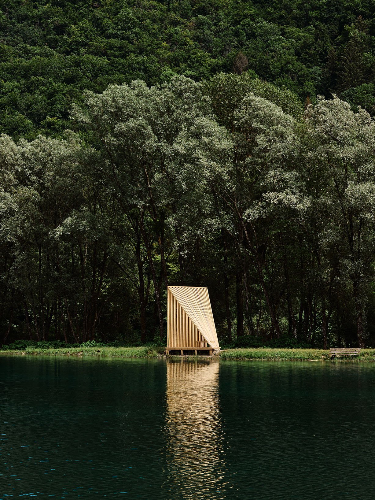 Slatted timber pavilion reflected in a mountain lake, framed by dense woodland in the background.