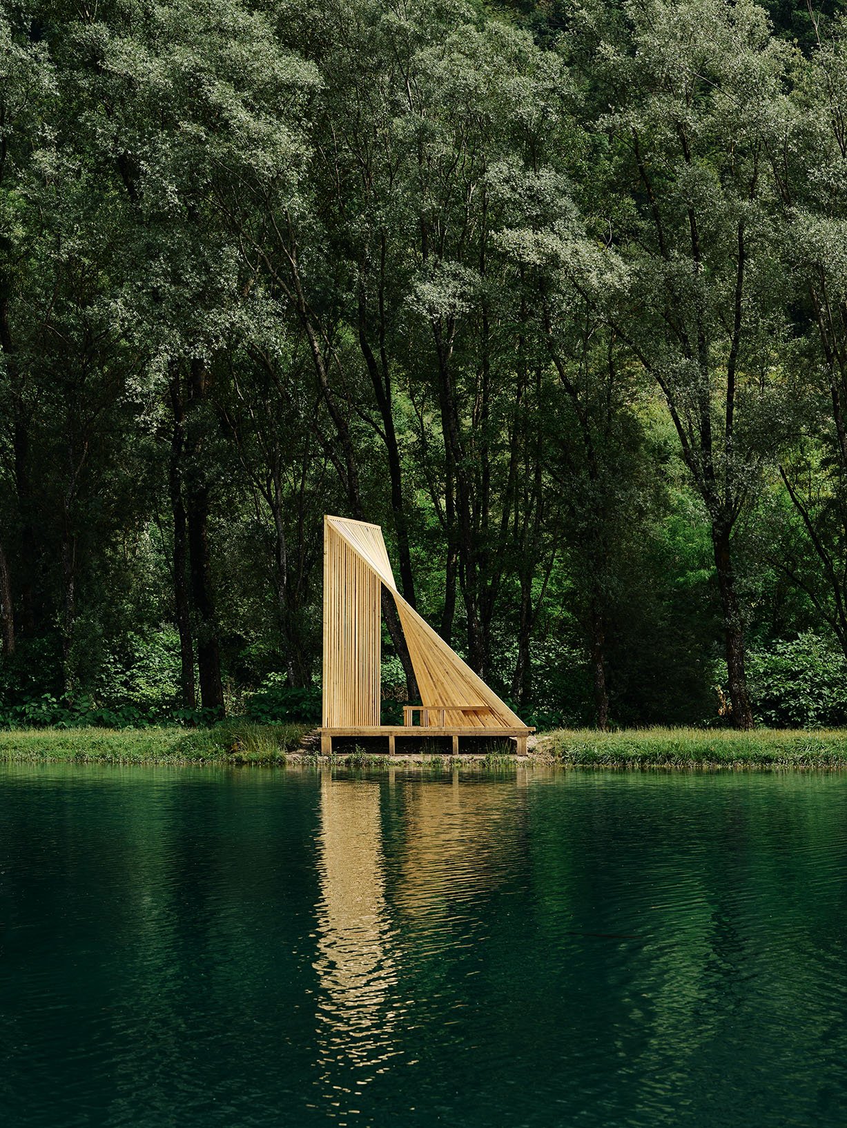 Sculptural timber pavilion composed of vertical slats forming a sail-like silhouette along the lakeshore.